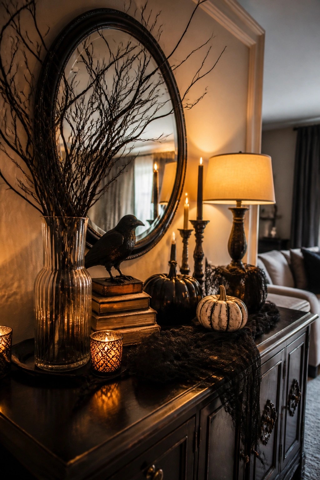 Dark wood console table with a black crow on stacked books, a glass vase holding bare branches, black pumpkins, and lit candles arranged in front of a round mirror.