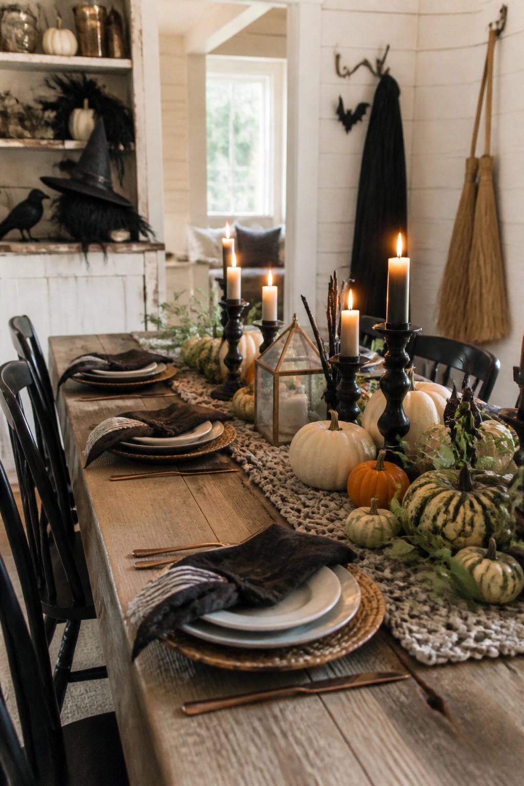 A farmhouse dining table styled with black candlesticks, lit white candles, assorted pumpkins, and dark place settings for Halloween.