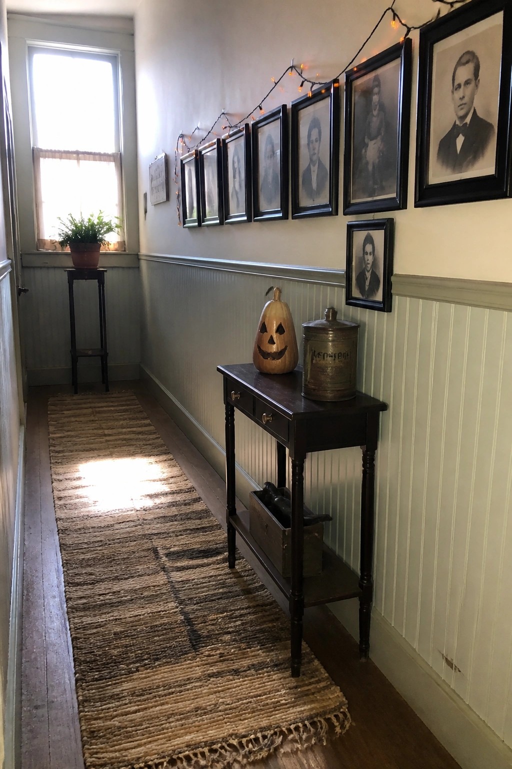 A narrow hallway with beadboard walls displays a long row of black-framed vintage portraits above a dark wooden console table that holds a carved pumpkin and a metal tin.