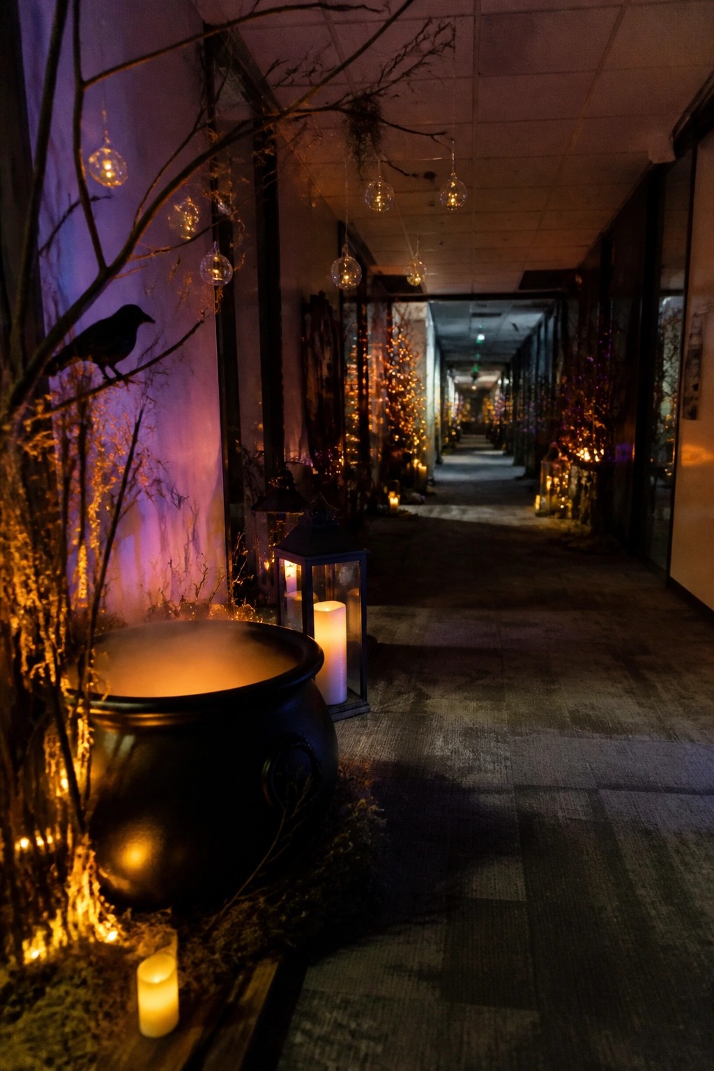 Long office hallway decorated for Halloween with a large black cauldron emitting mist, black lanterns, hanging glass orbs, and bare branches with crows along the walls.