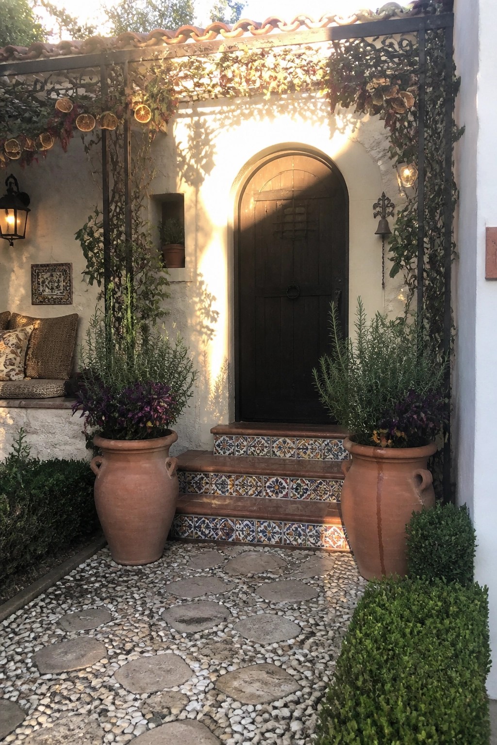 A wooden arched front door sits beneath a metal pergola with vines and dried citrus, flanked by two large terracotta pots on tiled steps.