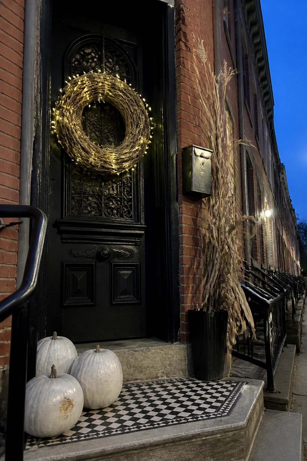 A black front door on a brick building features a large grapevine wreath wrapped in warm string lights, with three white pumpkins on a black and white checkered mat at the base.
