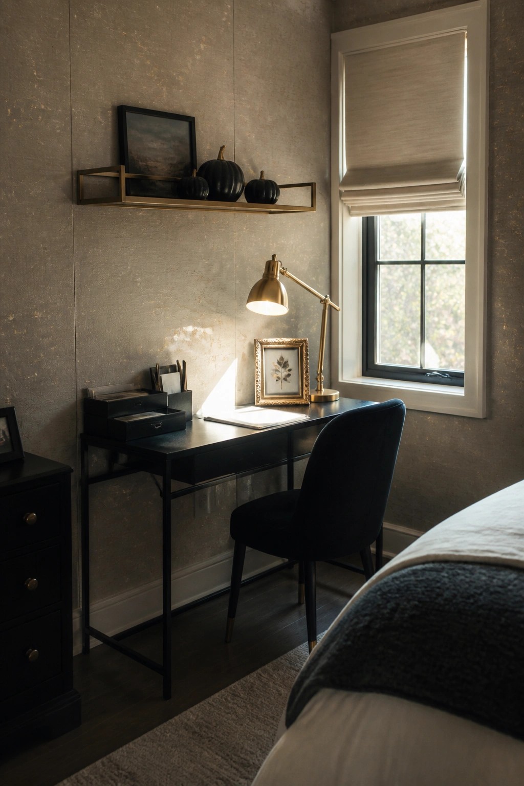 A black desk with a lit gold lamp, black pumpkins on a brass wall shelf, and a framed print beside the window in a room with textured walls.