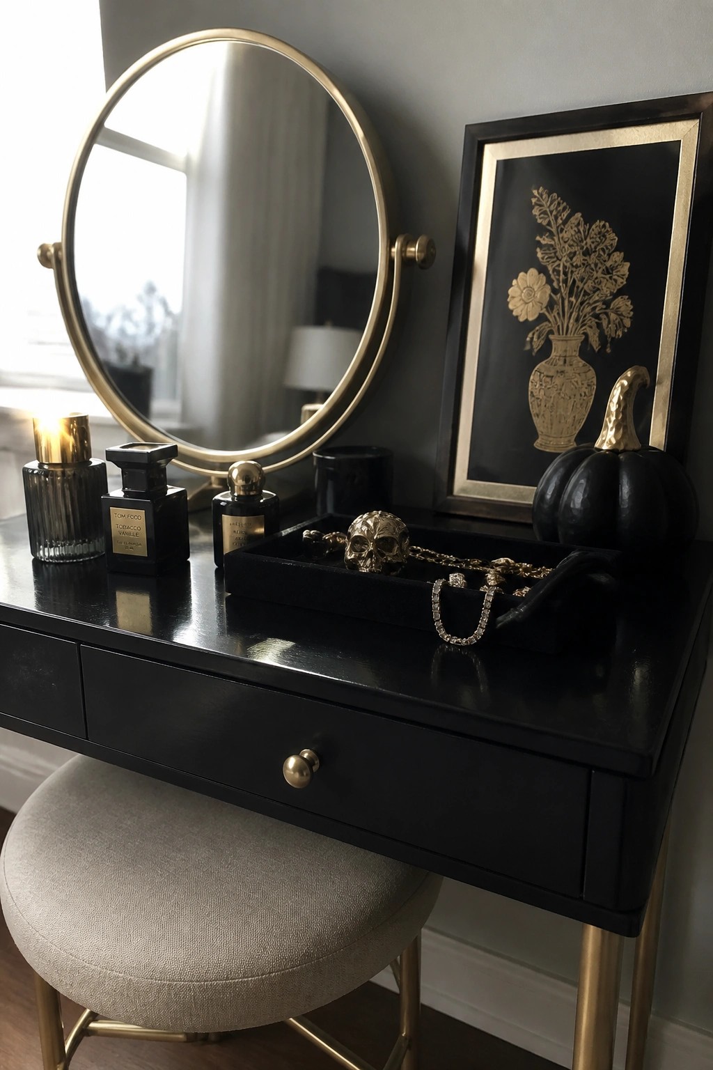 A black vanity table with round gold mirror, three perfume bottles, black jewelry tray holding a gold skull and chains, black pumpkin, and gold-framed floral artwork on a gray wall.
