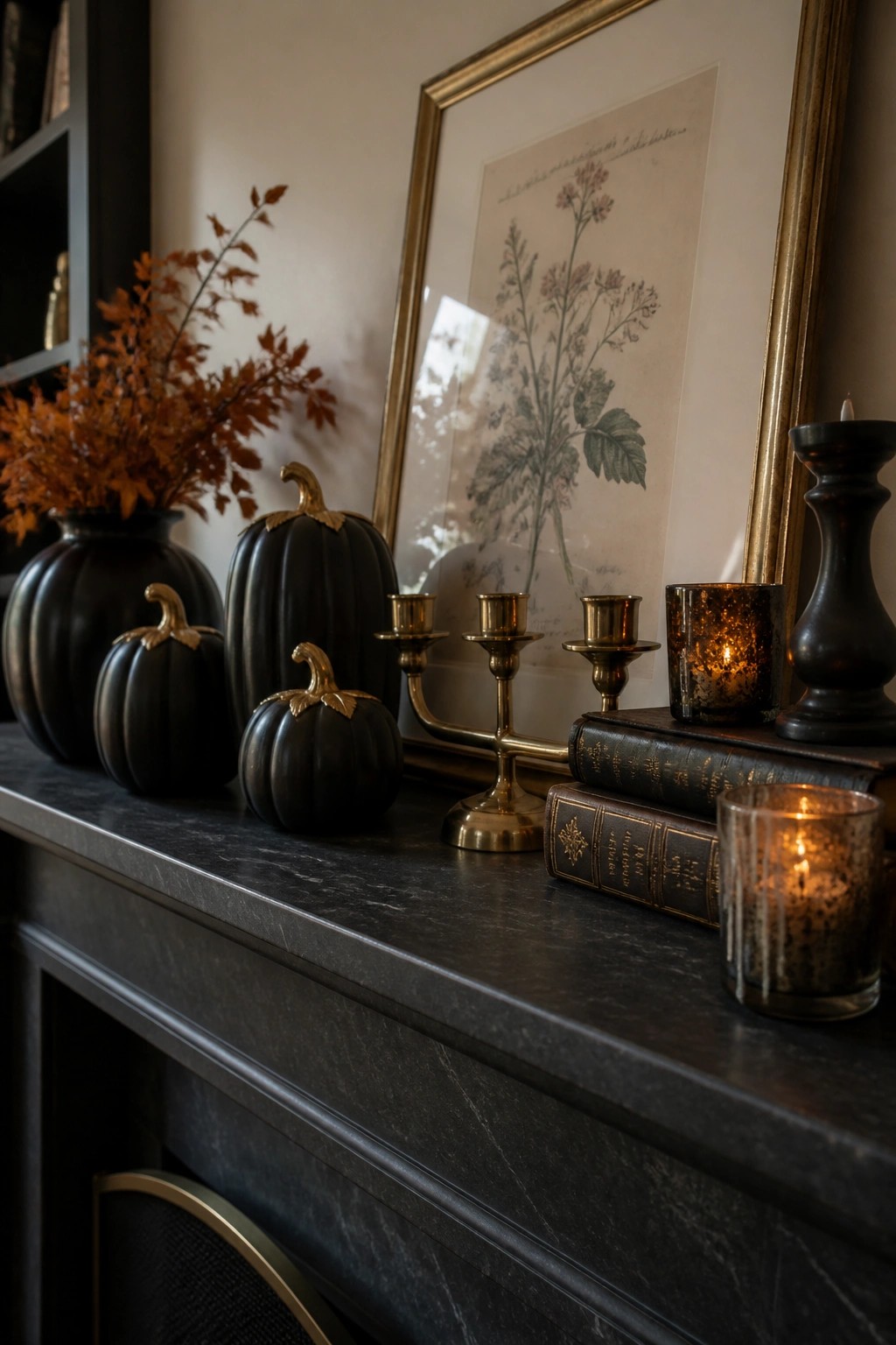 A dark marble mantel holds several black pumpkins with gold stems, brass candlesticks, lit candles in textured glass holders, and two stacked vintage books in front of a framed botanical print.