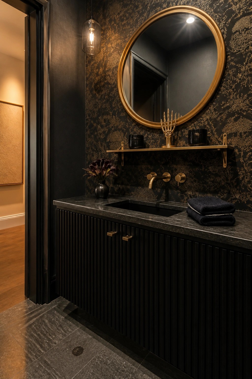 A black bathroom vanity with ribbed doors, black marble countertop, round gold mirror, and gold shelf mounted on dark wallpaper with a gold pattern.