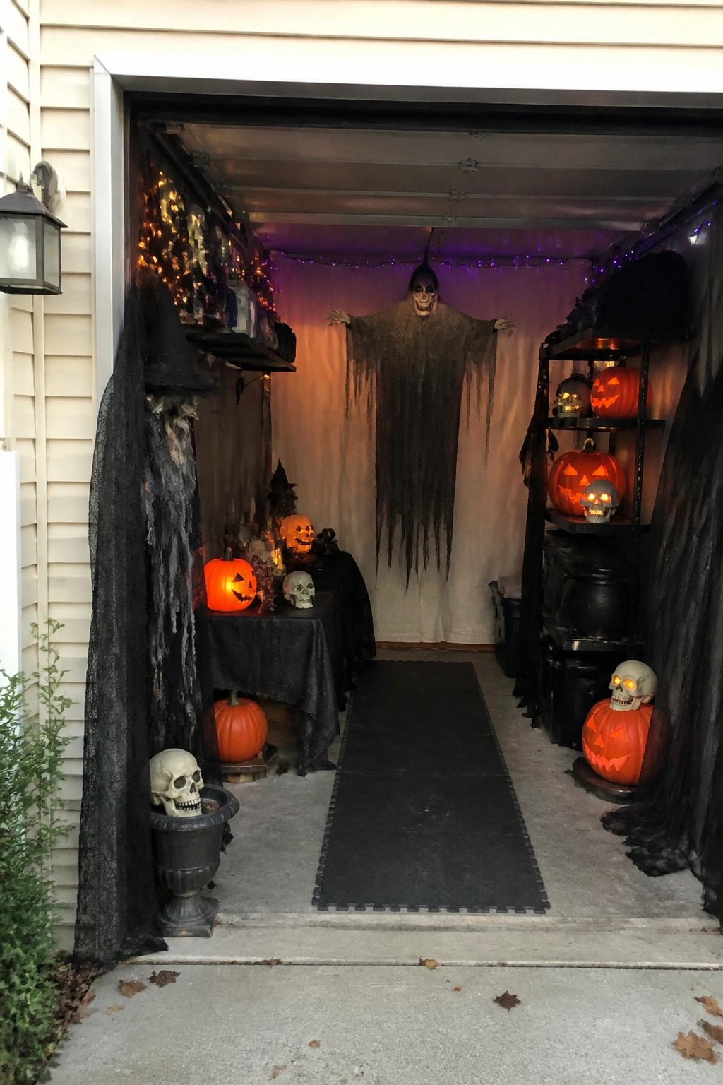 Open garage decorated for Halloween with a large hanging ghost figure at the back, multiple glowing jack-o'-lanterns on shelves, and skulls placed around the space.