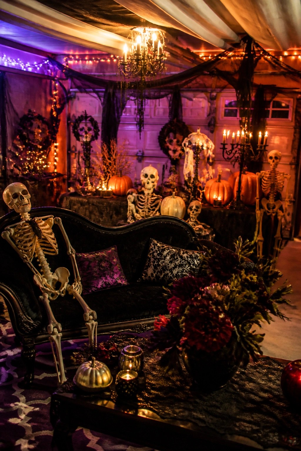 Garage party space with multiple skeletons posed on and around a black velvet sofa, surrounded by pumpkins, candles, and string lights.