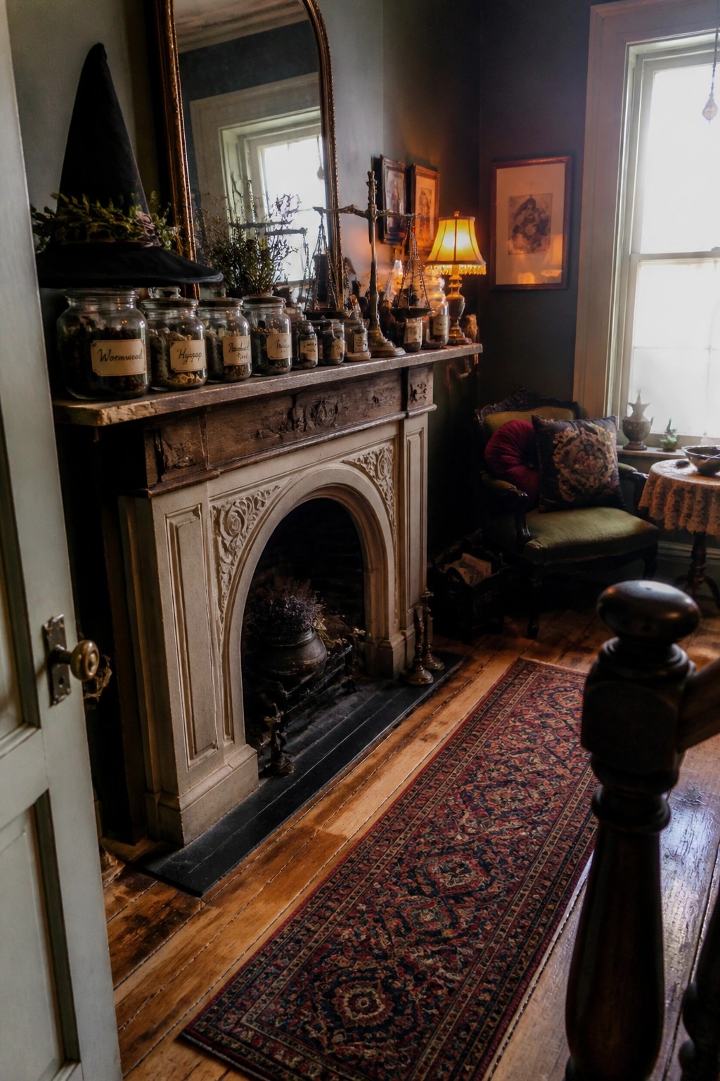 A fireplace mantel holds a row of glass herb jars with handwritten labels and a black witch hat resting on the left side.