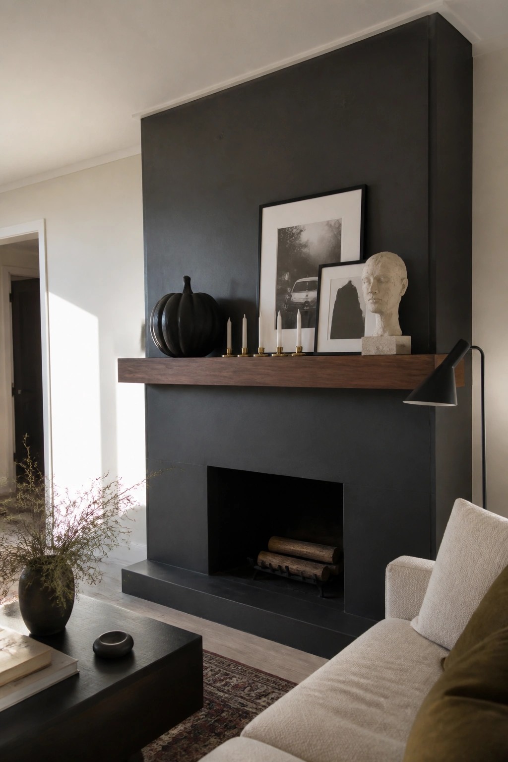 A dark painted fireplace mantel with a black pumpkin, white taper candles, framed black and white photos, and a stone bust sculpture.