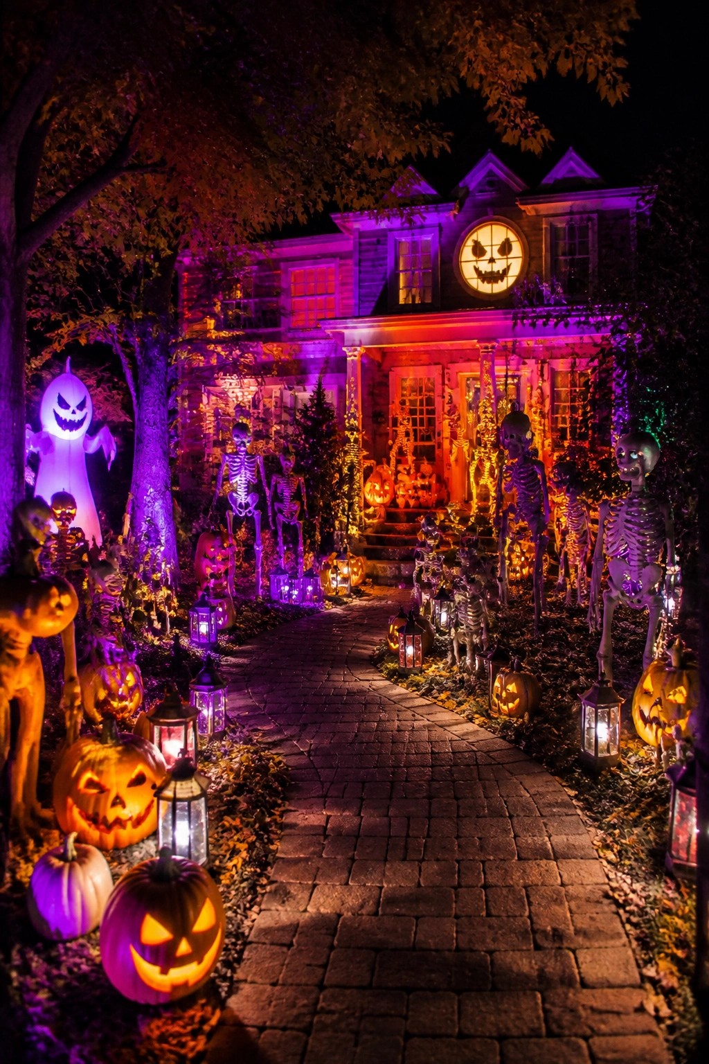 Brick walkway lined with standing skeletons, glowing jack o lanterns, and lanterns leading to a purple lit house porch.