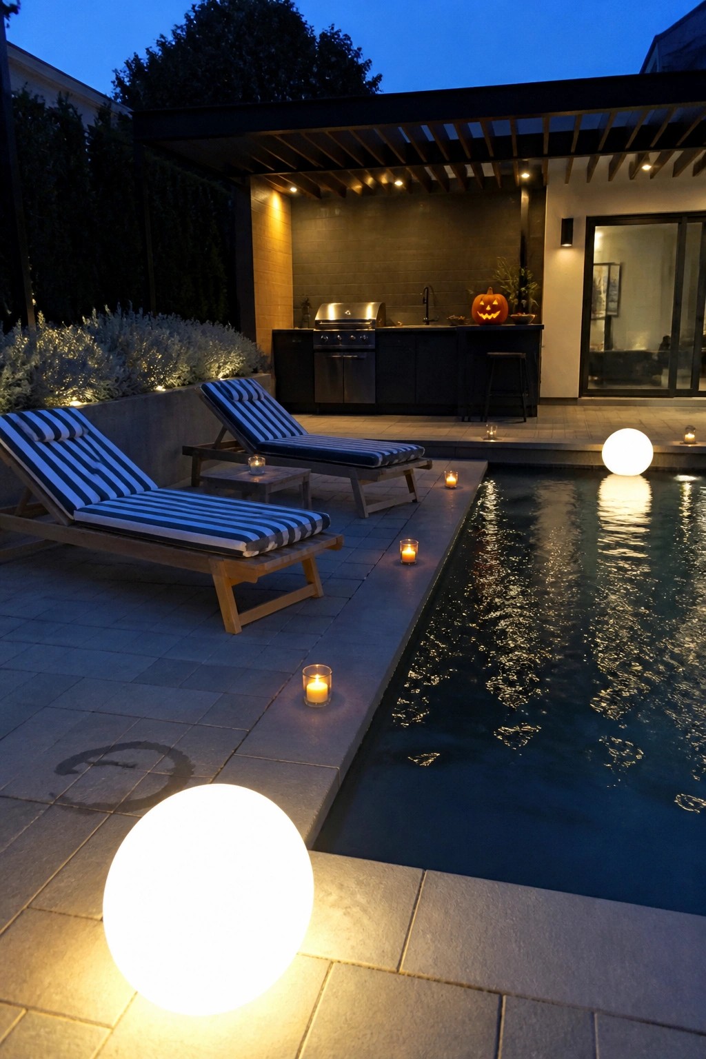 Nighttime view of a backyard pool with two large glowing white orbs on the stone deck, striped lounge chairs, an outdoor kitchen, and small candles along the pool edge.