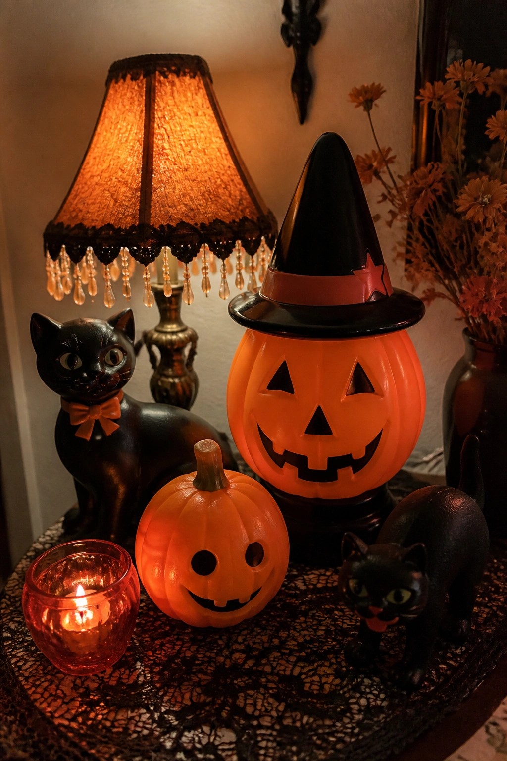 A tabletop Halloween vignette with a large jack o lantern wearing a witch hat, a smaller carved pumpkin, two black cat figurines, and a lit red candle on a lace cloth.