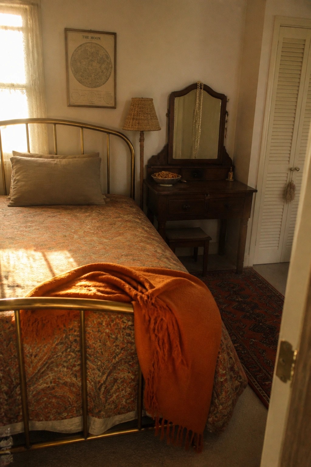 A bedroom interior showing a brass bed with a patterned quilt and orange throw, a wooden vanity with mirror, and a framed vintage moon chart hanging on the wall above a woven lamp.