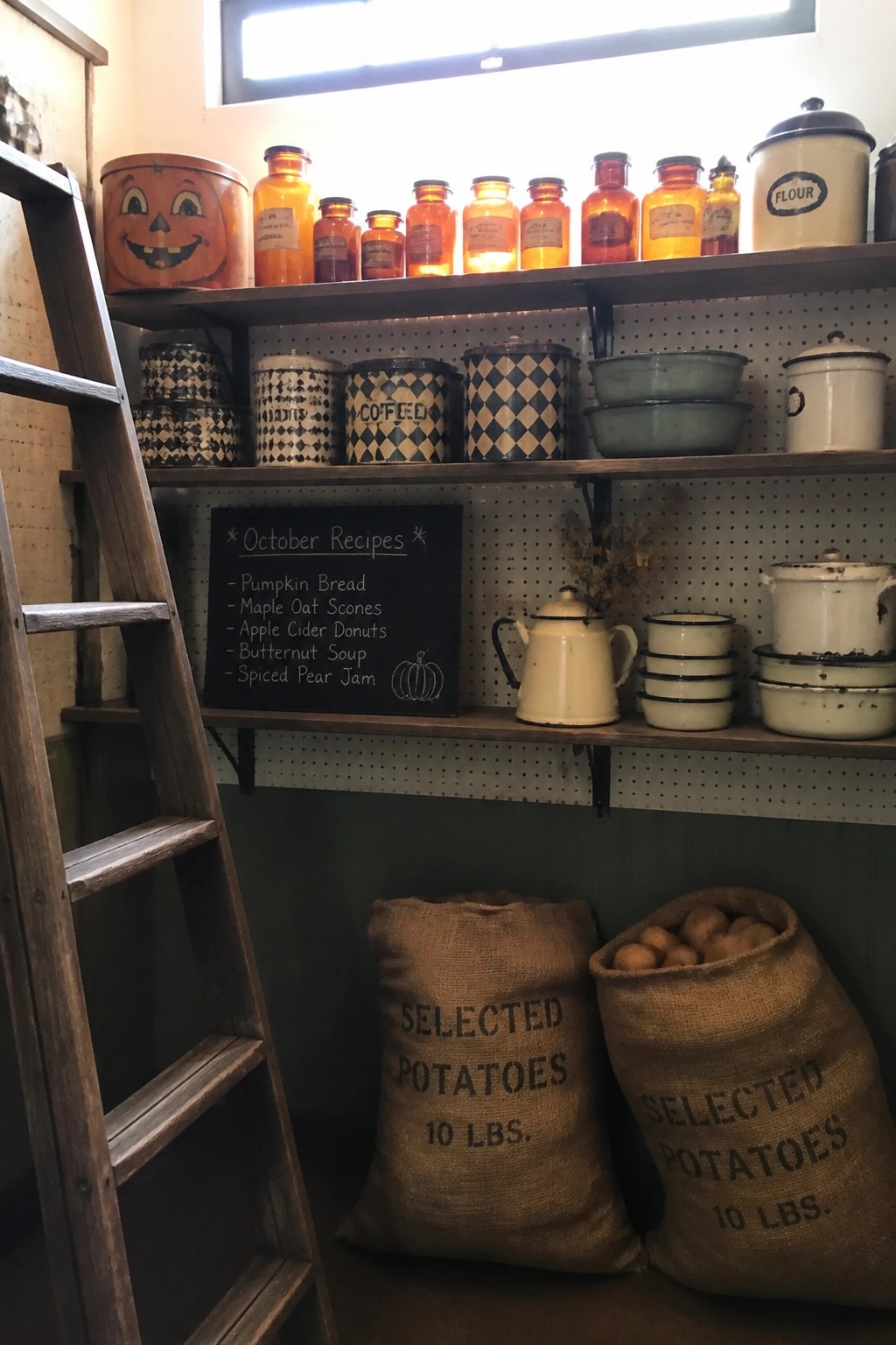 Wooden open shelves display vintage glass jars, patterned metal canisters, enamelware bowls, and a chalkboard listing October recipes, with two burlap potato sacks on the floor below.