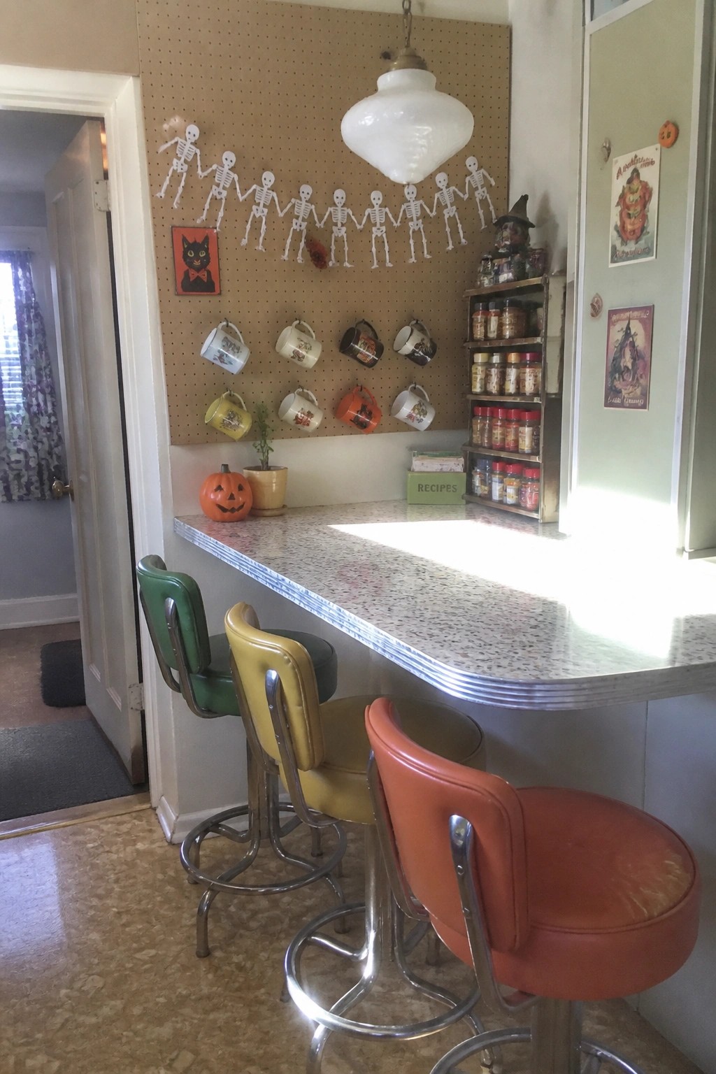 A retro kitchen counter with three stools faces a pegboard wall holding a skeleton garland, Halloween mugs, and small prints.