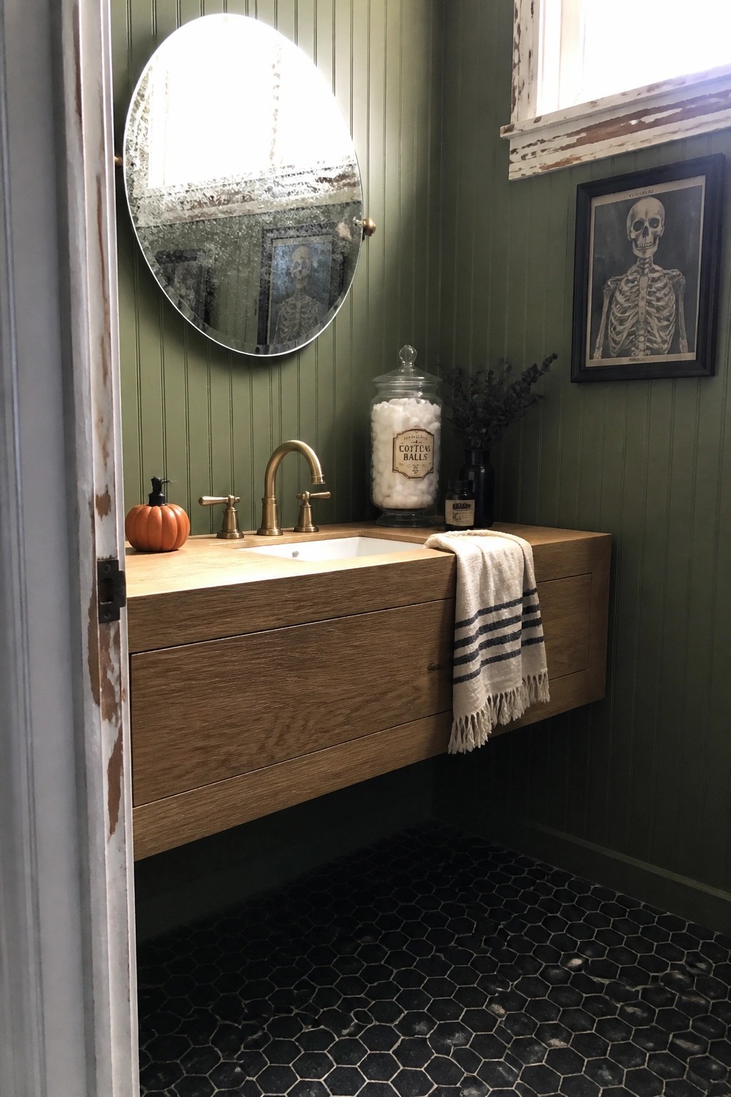 Green-walled bathroom with wooden vanity, round mirror, skeleton art, and black hex tiles.
