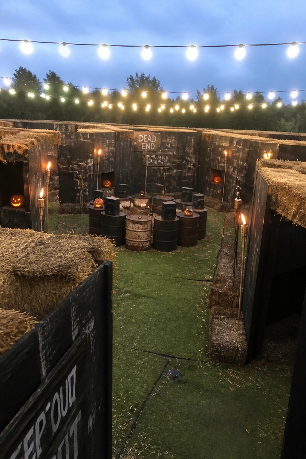 Twilight Halloween maze with hay bales, pumpkins, barrels, torches, and string lights.