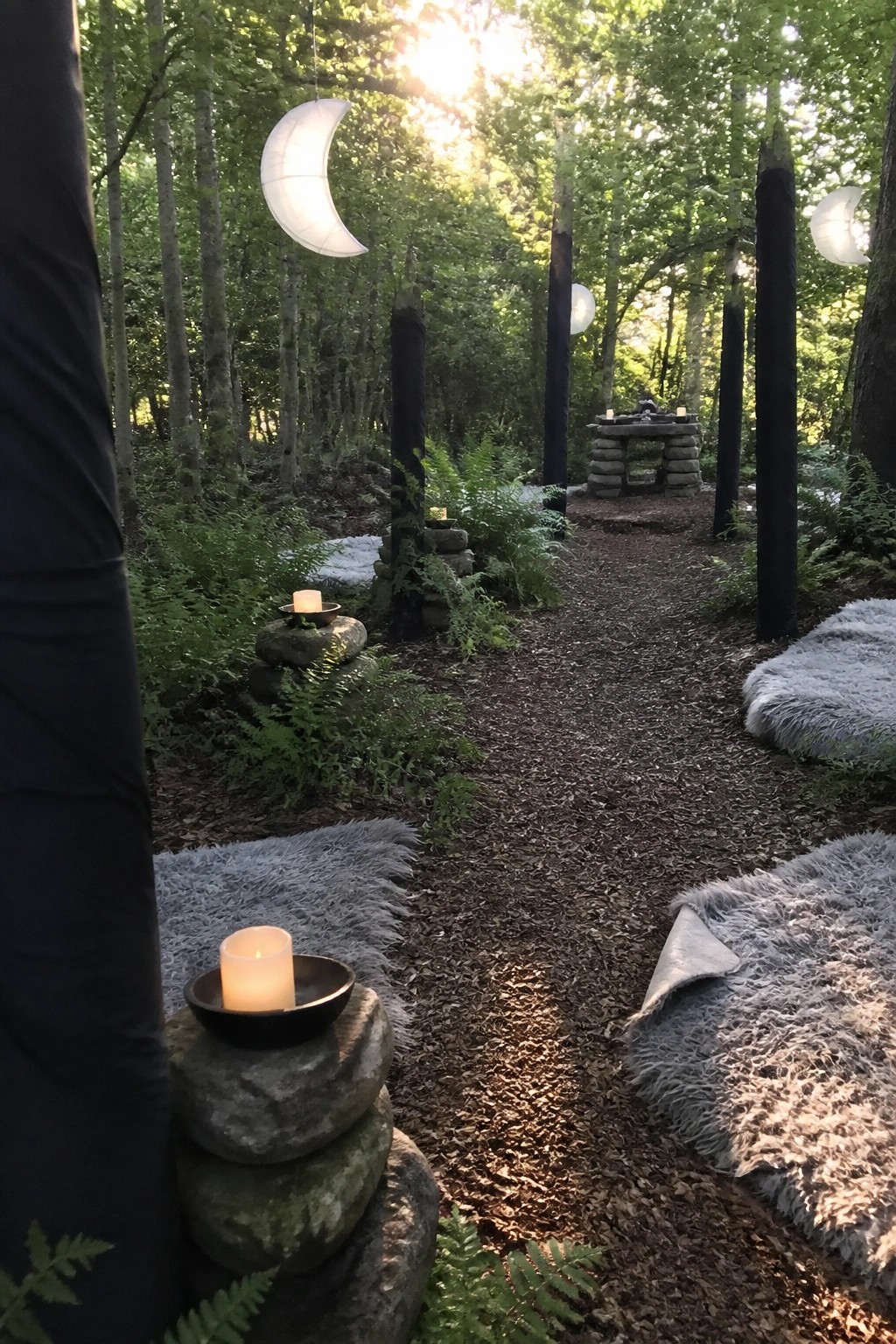 Forest path with hanging crescent moon lanterns, candles on rocks, and gray fur rugs.