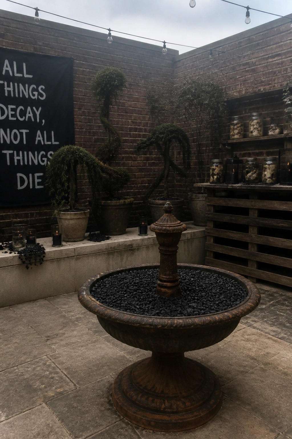 A large rusted fountain filled with black gravel stands in a brick courtyard with potted plants, candles, and a black banner on the wall.