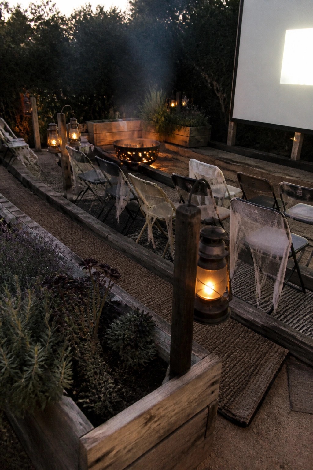 A backyard at dusk with a large projection screen, a fire pit, folding chairs decorated with spider webs, and several lit lanterns placed around wooden planters and rugs.