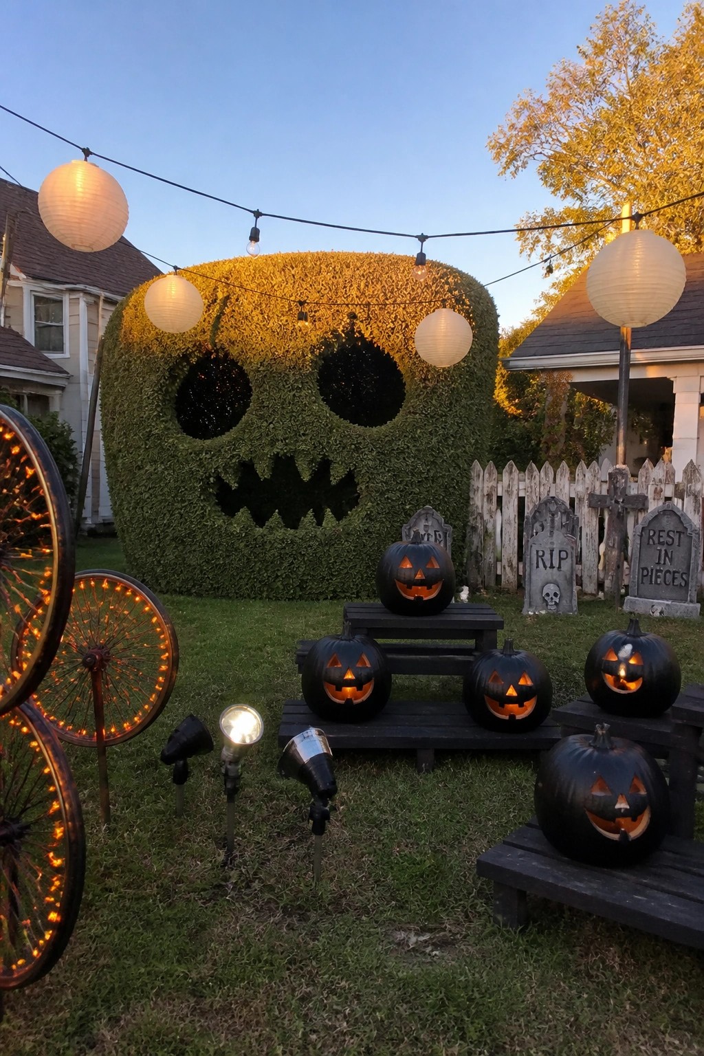 A large hedge trimmed into a skull shape with dark eye and mouth openings, lit from within, and surrounded by carved pumpkins and tombstones on a lawn.