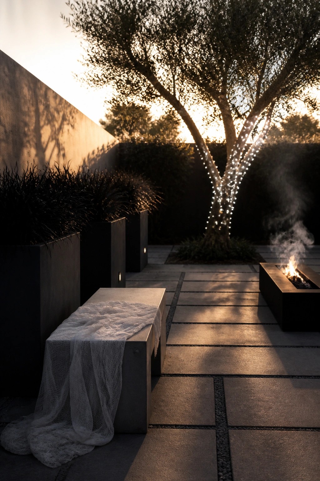 A modern patio at dusk features a tree wrapped in string lights beside a rectangular fire pit, with a concrete bench draped in white lace fabric in the foreground.