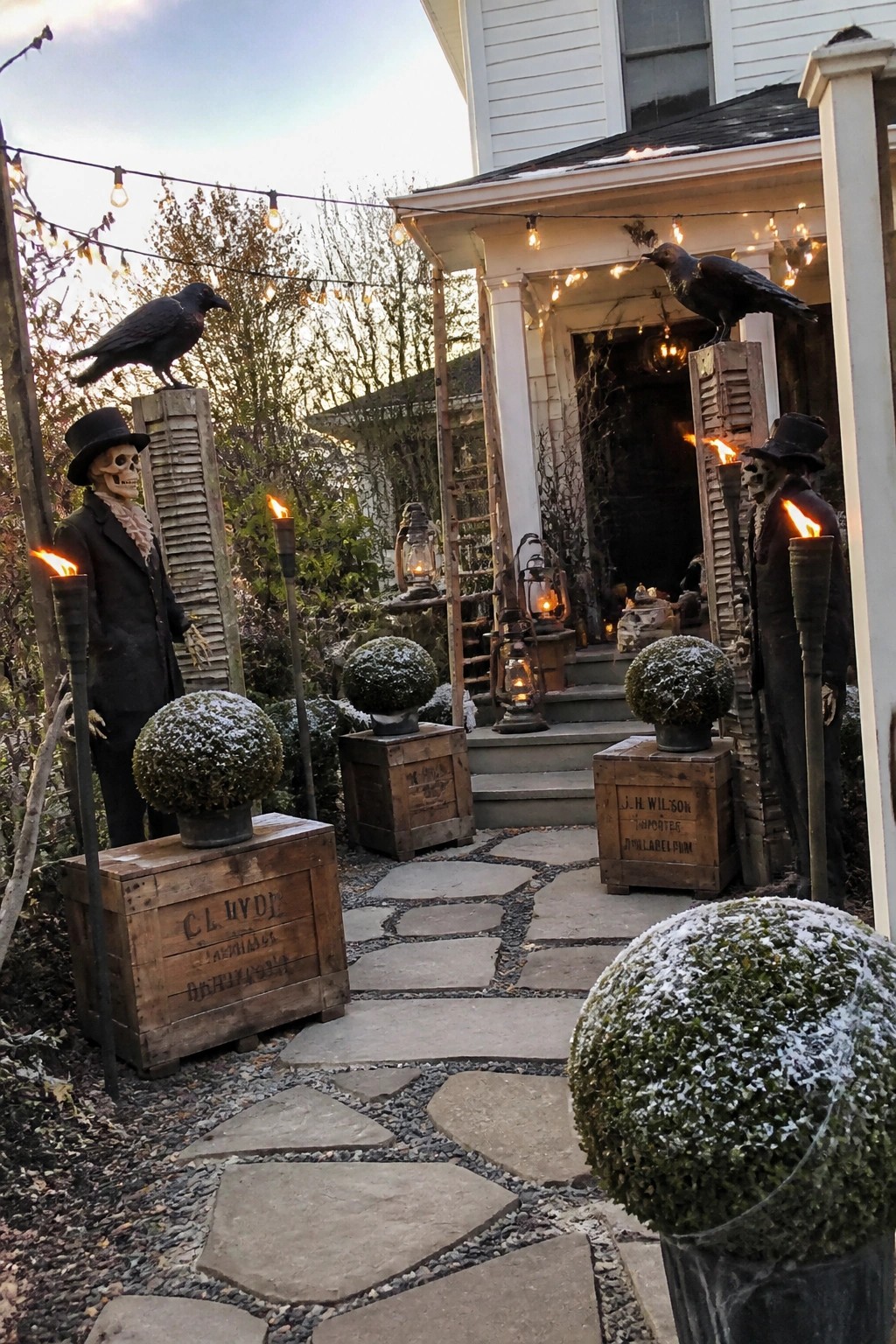 A front yard Halloween display with skeleton figures in top hats standing on wooden posts and crates, black crows, lit torches, and a stone path leading to a house porch.