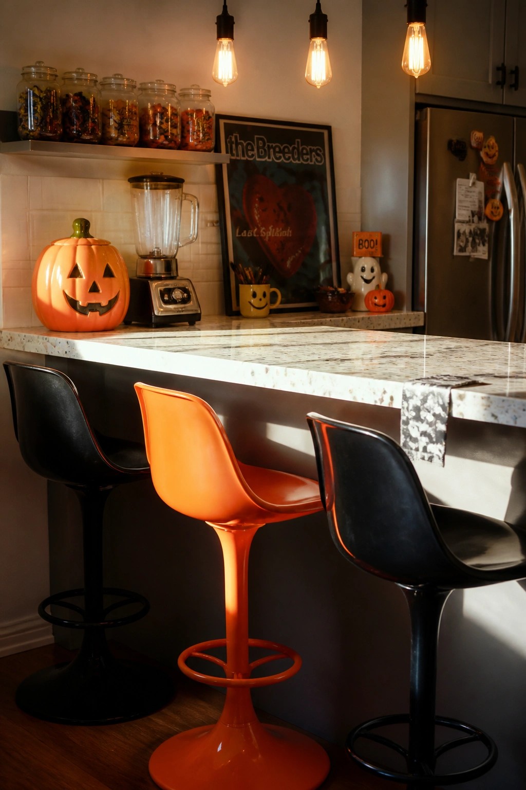 A kitchen counter area with three modern bar stools, one bright orange and two black, next to an orange pumpkin cookie jar and other small Halloween decorations.