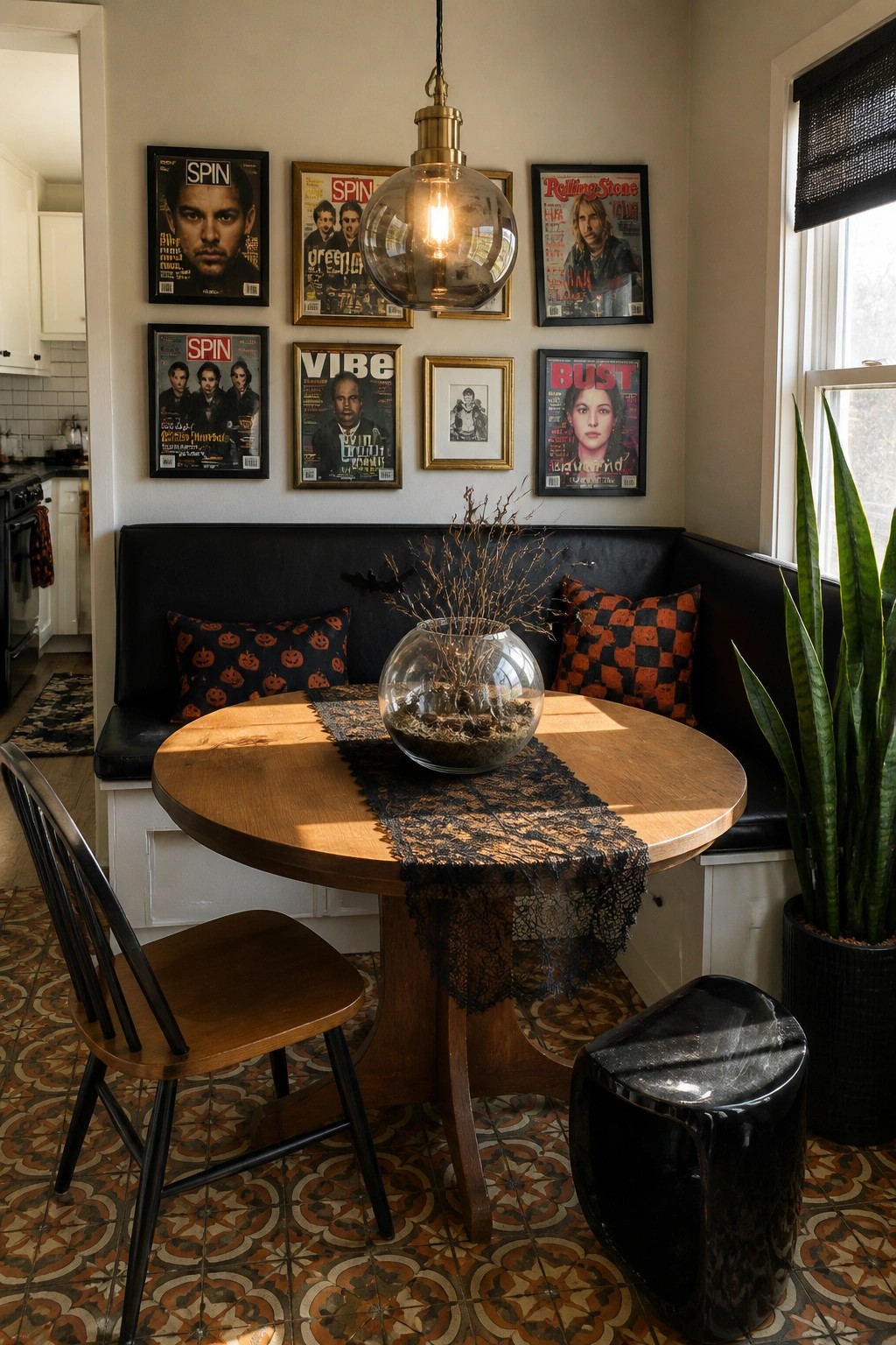 A round wooden dining table sits in front of a black built-in bench with pumpkin-patterned pillows, while framed 90s music magazines hang on the wall above.