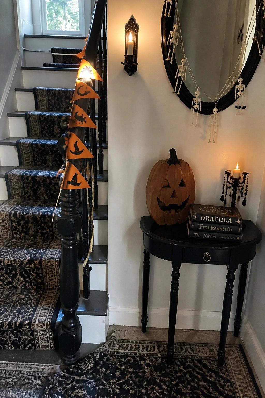 A wooden carved pumpkin sits on a black round table next to three stacked horror books, with a skeleton garland on a mirror above and a Halloween banner hanging on the nearby staircase.
