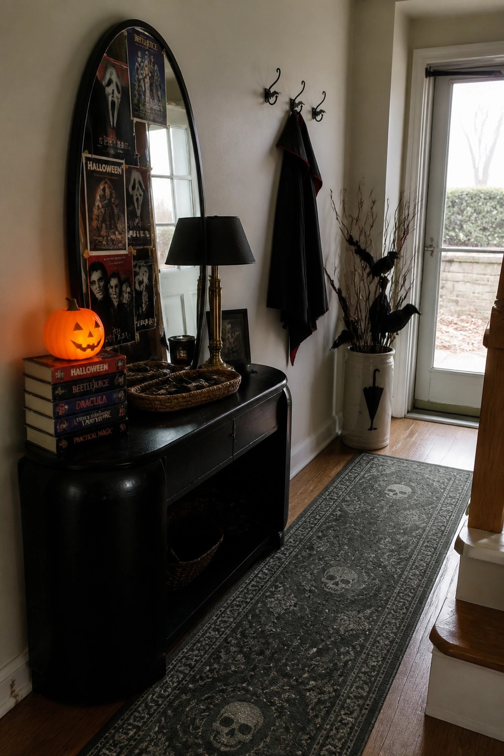 A black console table against a wall holds a stack of Halloween, Beetlejuice, and Dracula books topped with a lit jack-o'-lantern, with movie posters attached to an oval mirror above it and a dark skull-patterned rug on the floor.