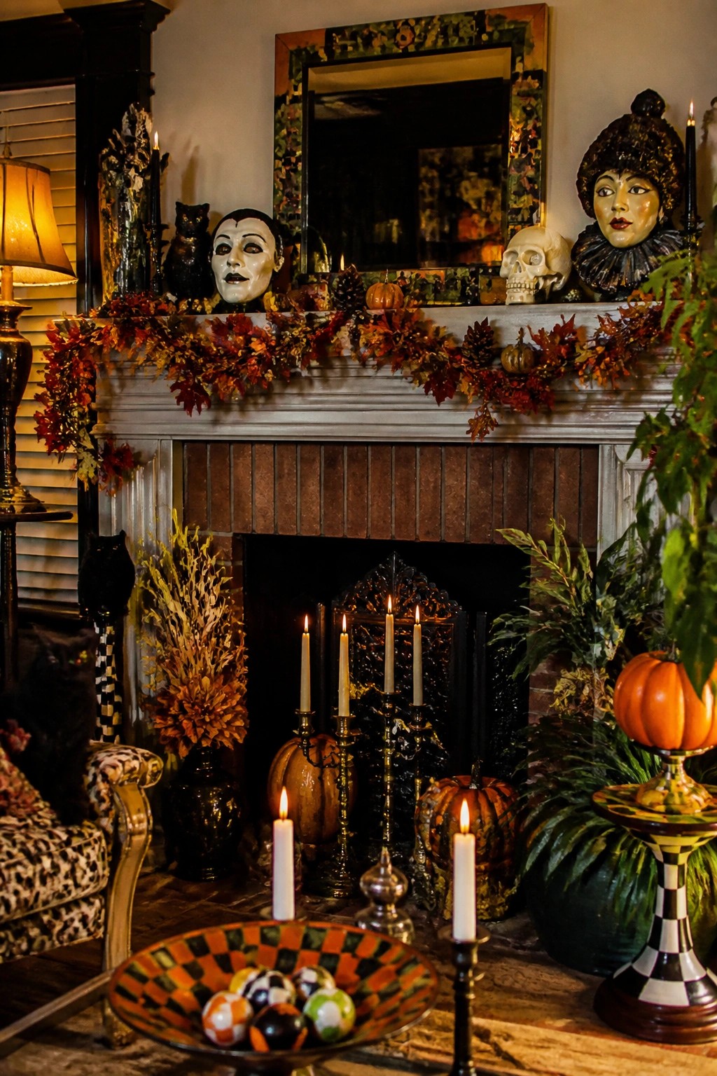 Fireplace mantel decorated for Halloween with a white theatrical mask, colorful harlequin bust, skull, black cats, candles, and autumn garland.