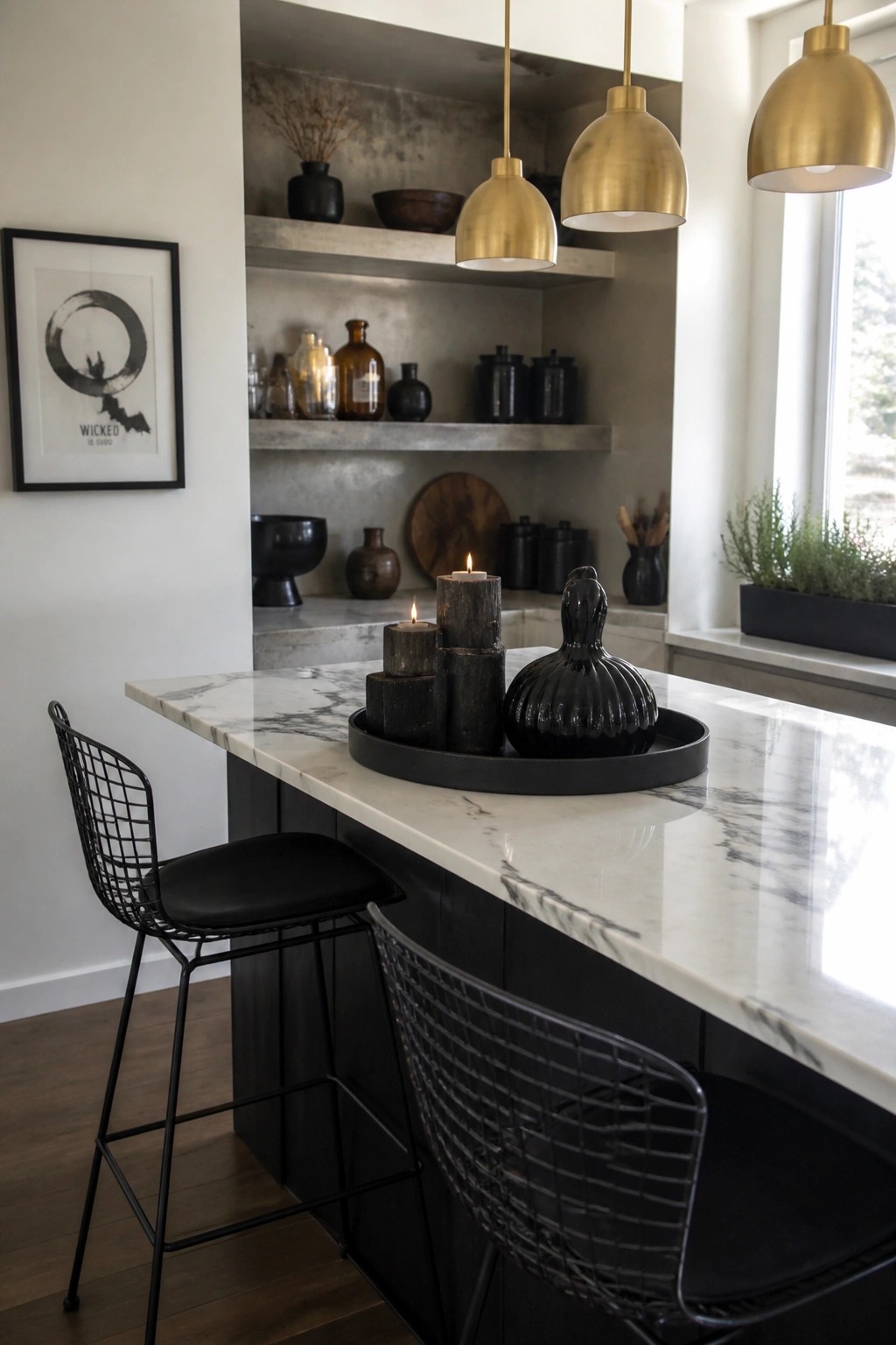 A kitchen island with white marble top, two black wire bar stools, and a black tray holding pillar candles on wood blocks beside a large black fluted vase, with gold pendant lights overhead.