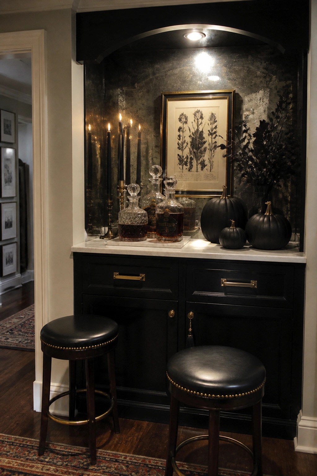 A black built-in bar cabinet with a white marble top displays glass decanters, lit black taper candles, a framed botanical print, and several black pumpkins, with two black leather stools placed in front.