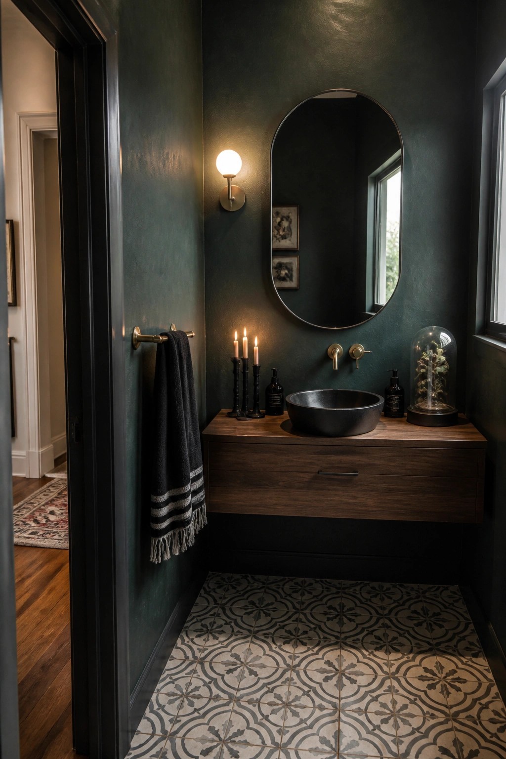 A bathroom with dark green walls, a floating wooden vanity, black vessel sink, oval mirror, and three lit candles on the counter.