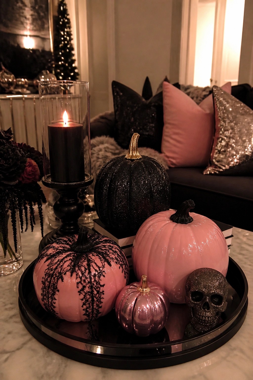 A round black tray holds pink pumpkins with black lace, a glitter skull, and a lit black candle on a marble surface.