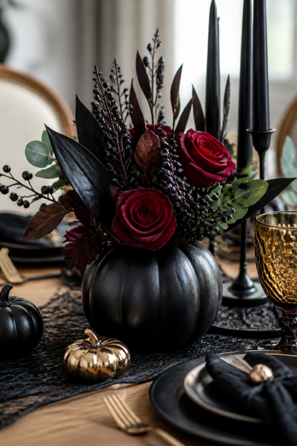 Black pumpkin vase holding red roses and dark foliage as the centerpiece on a dining table set with black plates, black candles, and small black and gold pumpkins.