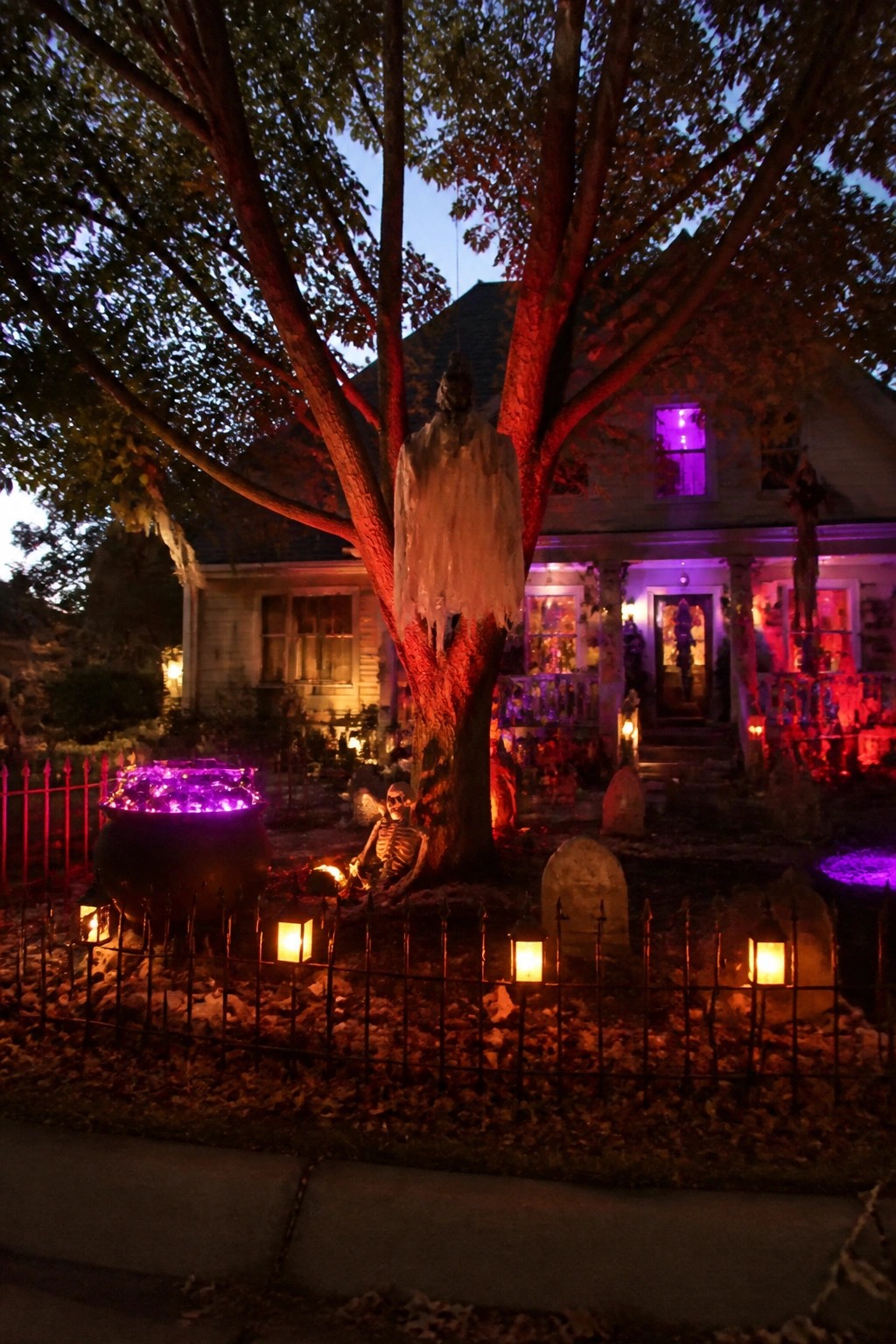 A front yard Halloween display with a large white ghost hanging from a tree, a skeleton seated below, a glowing purple cauldron, and several tombstones inside a low fence.