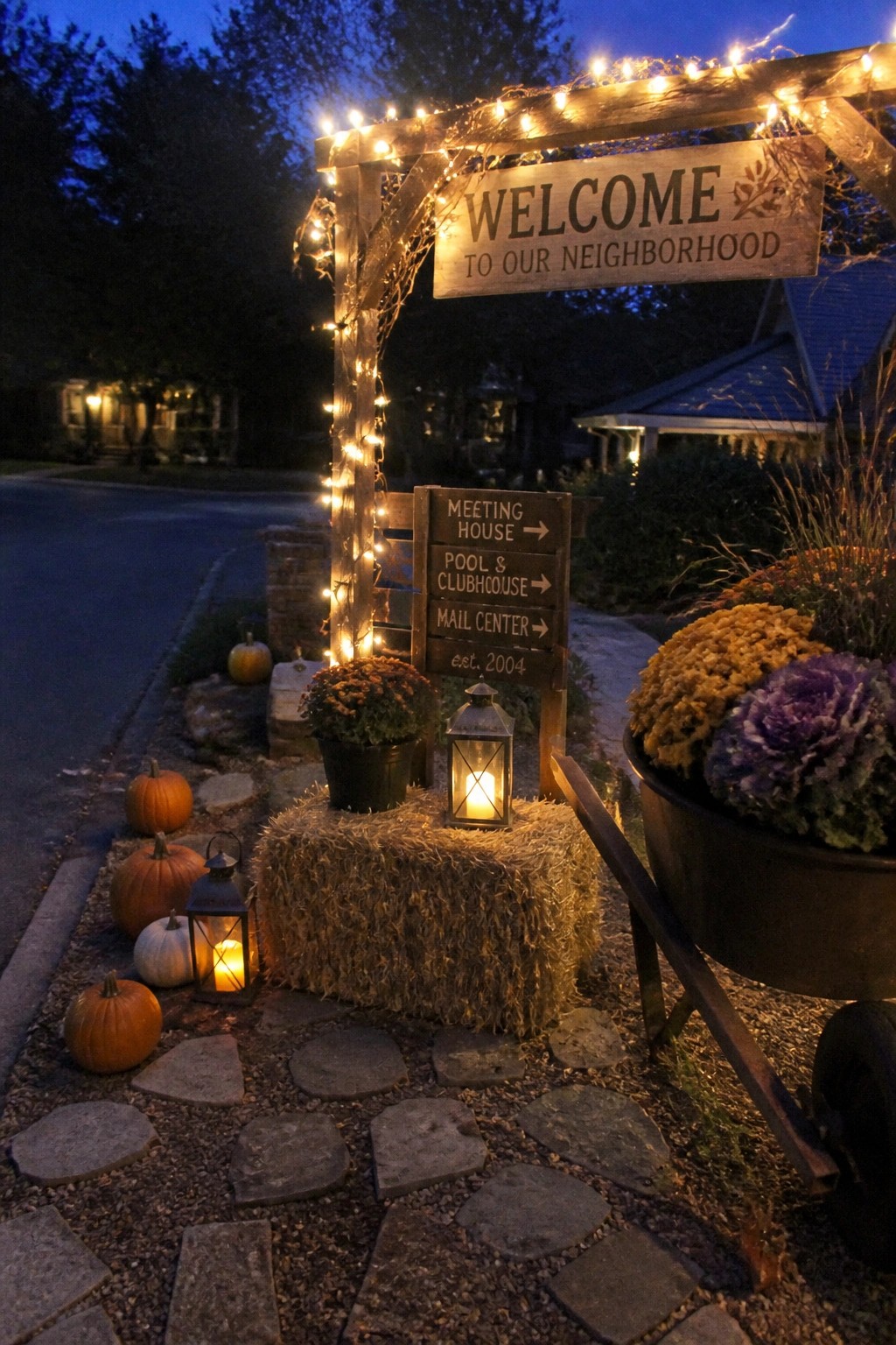 Autumn neighborhood entrance with lit wooden arch, pumpkins, hay bales, and signs at dusk.