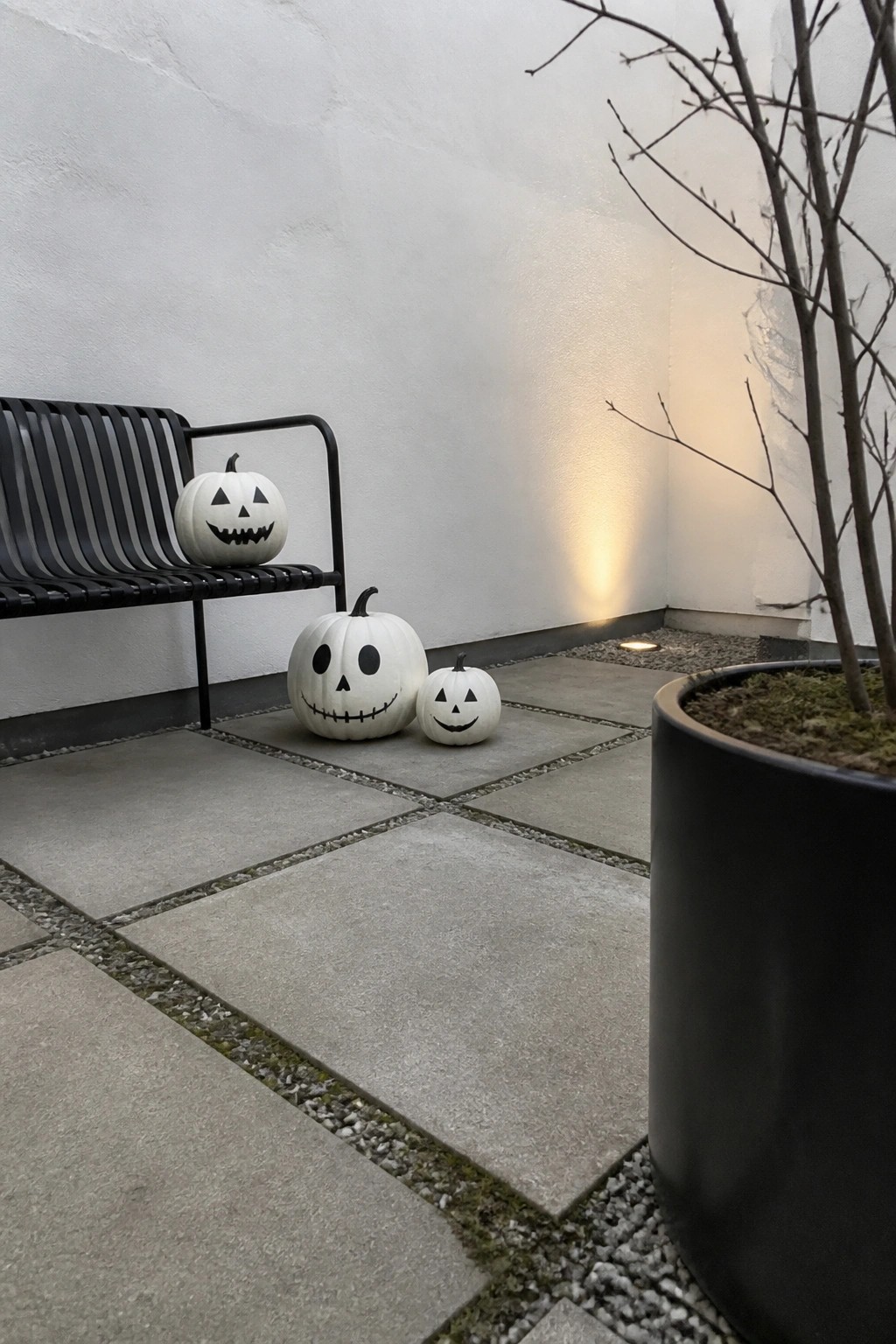 Three white carved pumpkins on gray tiles beside black bench and planter