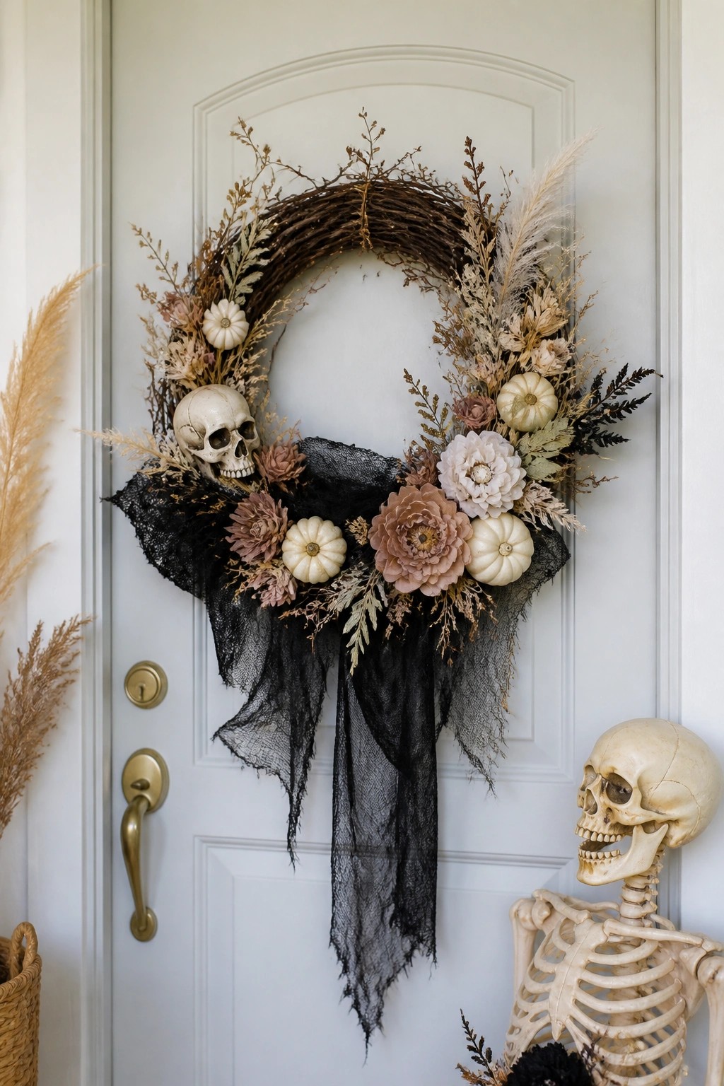 A white front door with a large wreath featuring a skull, black fabric draping, dried foliage, and small white pumpkins, next to a standing skeleton decoration.
