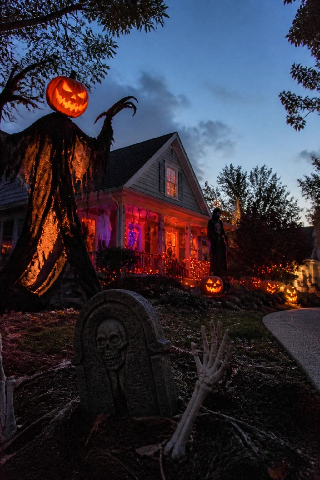Illuminated jack-o'-lantern scarecrow, skull tombstone, and skeletal hand in Halloween yard at dusk